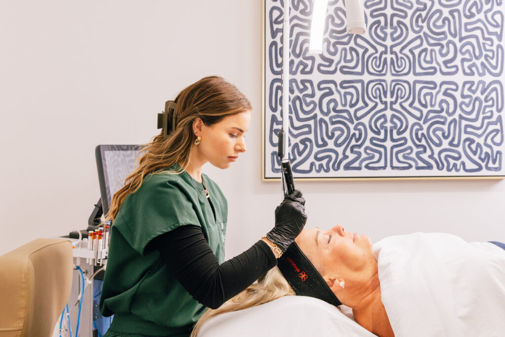 A provider at The Pip Rx administers a microneedling treatment in Overland Park. Wearing surgical gloves, she carefully passes the handheld microneedling device over her patient's forehead while her patient relaxes on her back.